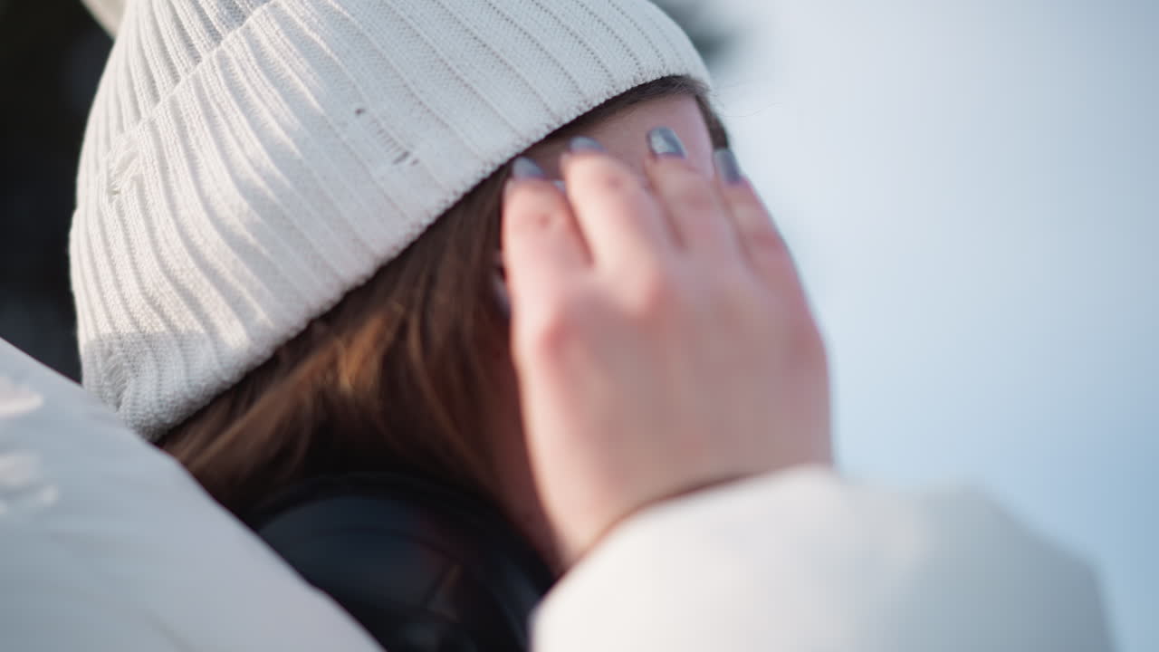 Close up side view of performer adjusting tinted white goggles while grooving to music under bright sky with wind tousling hair and manicured nails visible against soft winter coat and knit beanie