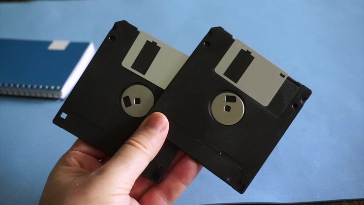 Mano maschile che tiene due floppy disk obsoleti da 3,5 pollici sopra una scrivania blu con un quaderno.