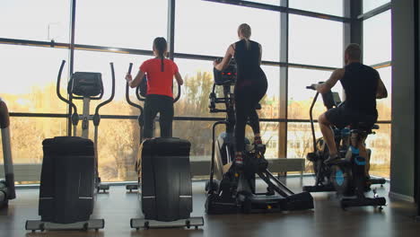 the view from the back: in the cardio area of the gym women on elliptical machines and a man on an exercise bike train near a large panoramic window in slow motion.