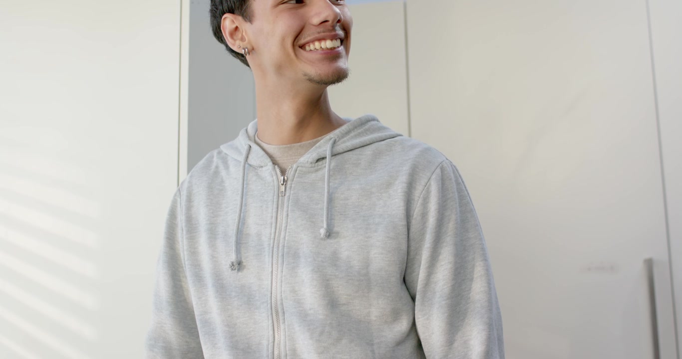 Uomo con felpa grigia con cappuccio che appoggia la mano sul bancone vicino a una finestra con tende, una pianta, girandosi a sinistra e sorridendo