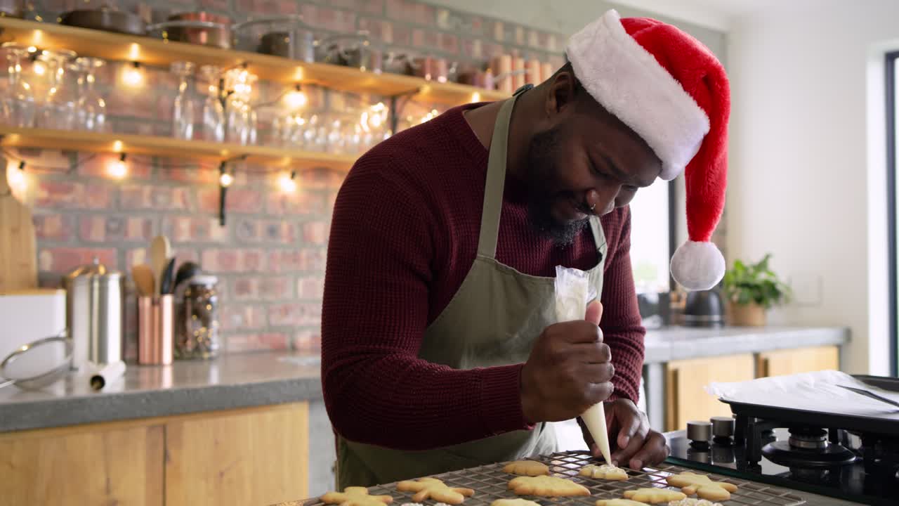African American man wearing Santa hat piping icing after baking cookies on cooling rack in kitchen