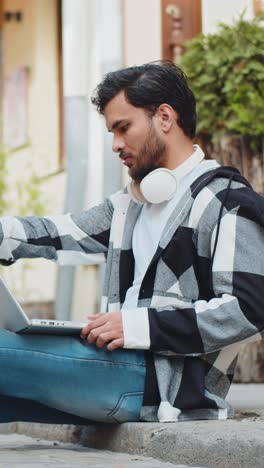 indian man freelancer sitting on street closes laptop screen finishing work smiling in city outdoors