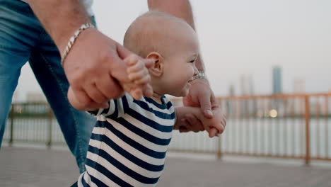 feliz bebé lindo aprende a caminar y da sus primeros pasos en la ciudad bajo el control de su padre sosteniendo las manos del bebé en el fondo de la ciudad