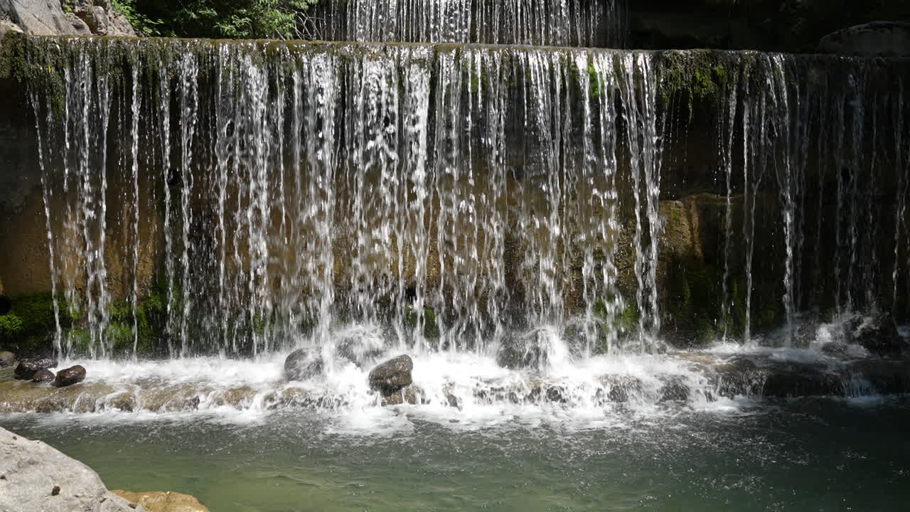 A slow-motion shot captures water cascading down a small, tiered fall surrounded by lush greenery near Walensee, Schweiz. Concept: Contemplative flow and engineered nature