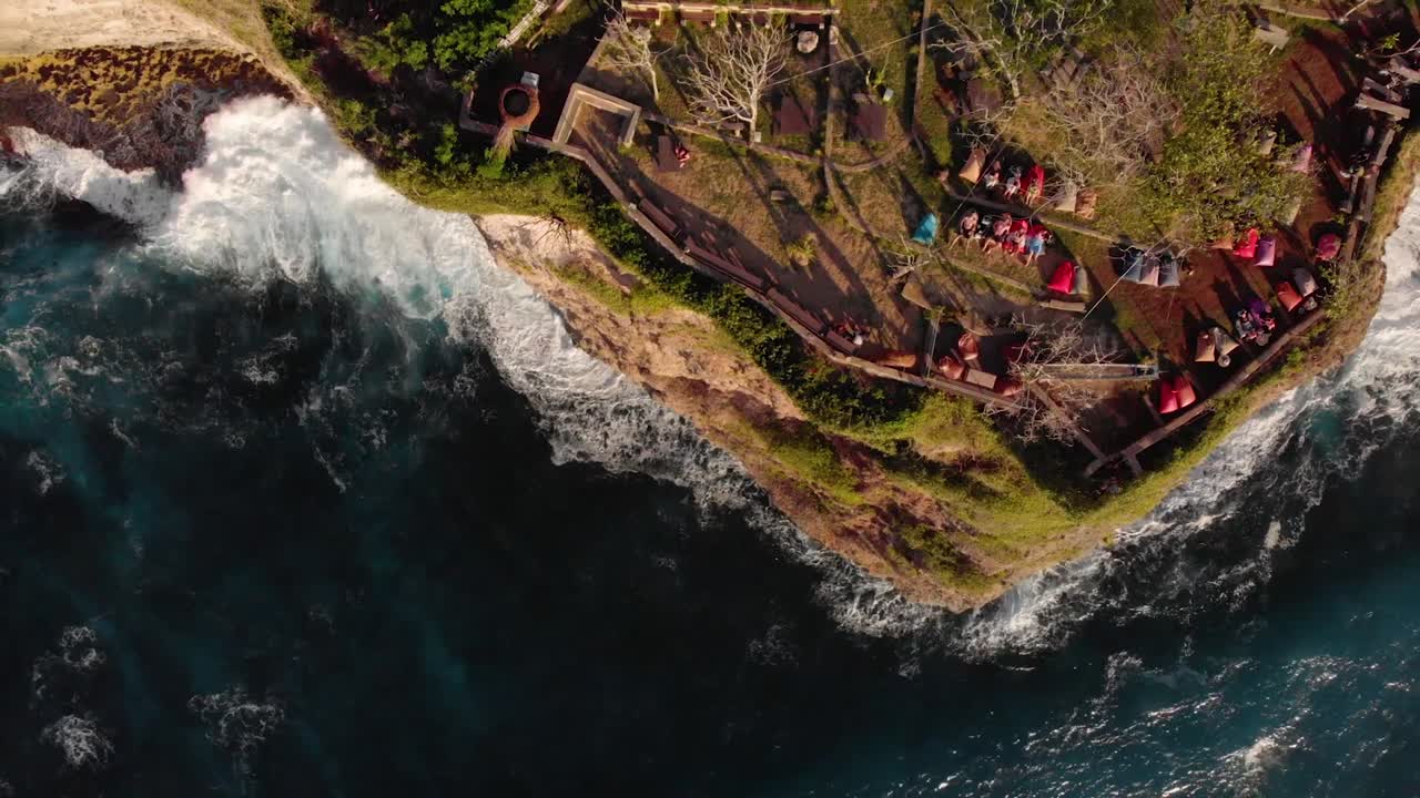 tiro de ojo de pájaro con drones del bar de la playa junto al acantilado con olas rompiendo debajo al atardecer en bali