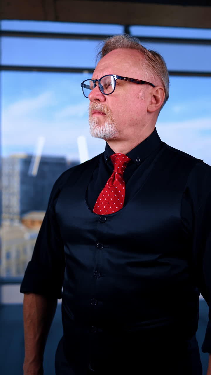 Confident Caucasian male in black shirt, vest and red tie checks his eyeglasses. Portrait of a businessman in office. Vertical video.