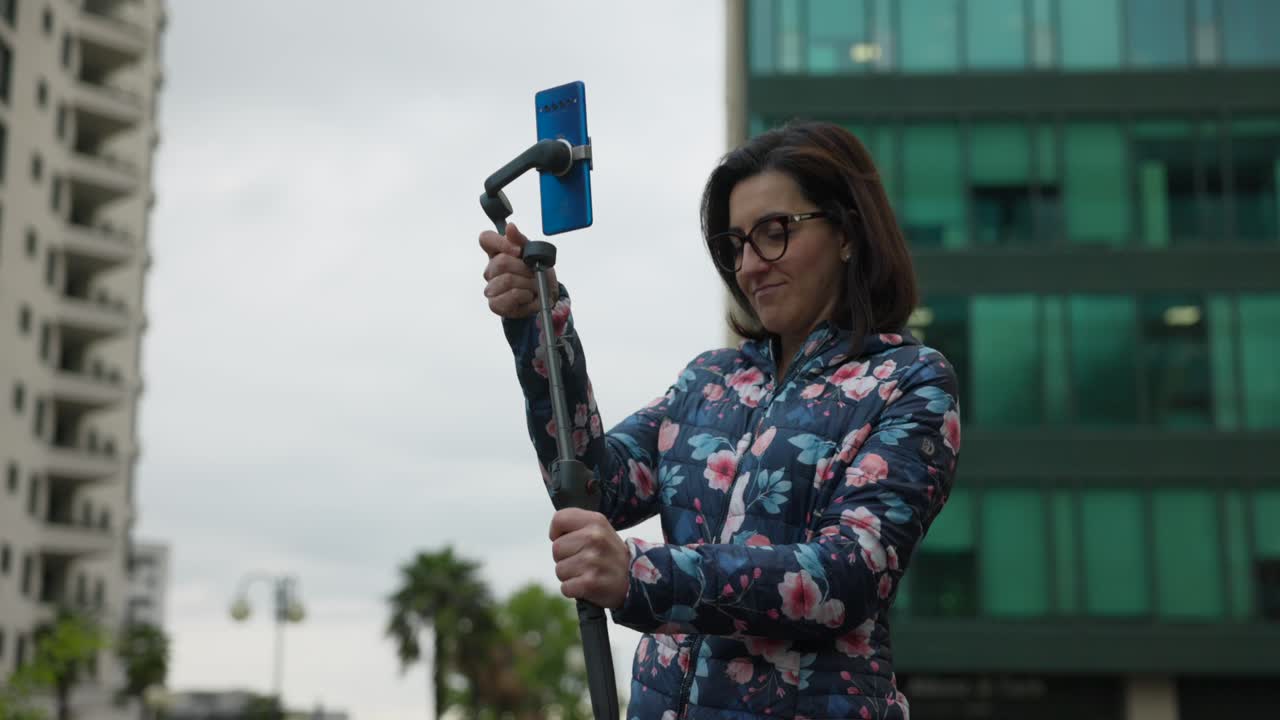 Brunette Caucasian woman with glasses holds and extend telescopic gimbal stabilizer with mobile phone