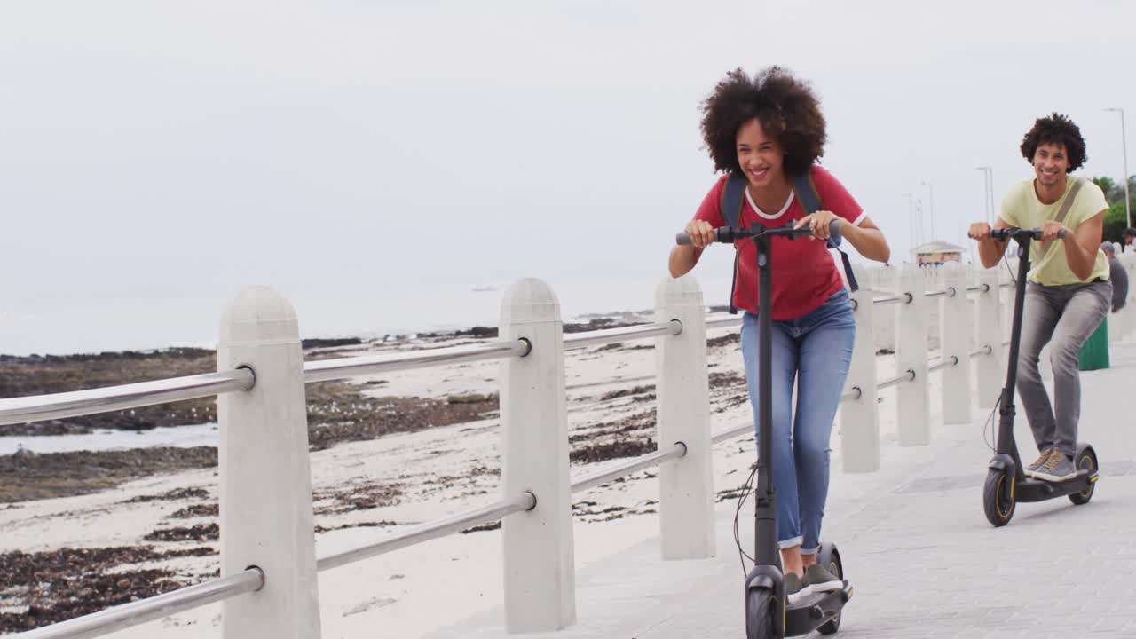 афроамериканская пара улыбается во время вождения скутеров вместе на набережной возле пляжа