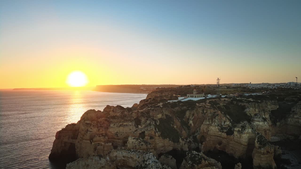 A câmera se afasta das espetaculares falésias costeiras e do farol da Ponta da Piedade, revelando rochas banhadas pelo sol, águas cintilantes e um pôr do sol vibrante ao longo da costa do Algarve
