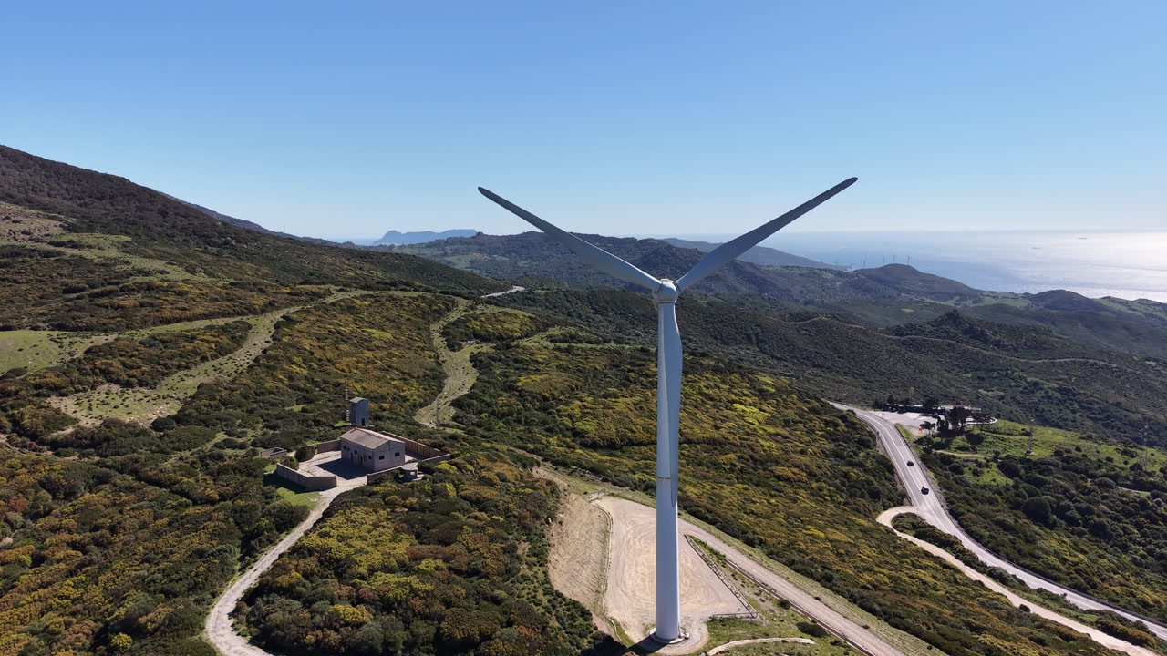 Aerial orbit around a wind turbine on a mountain ridge with a view of the Gibraltar strait in the Horizon.