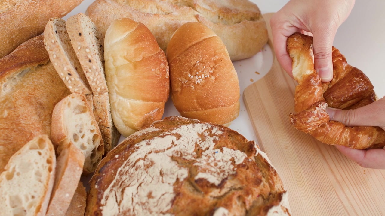 Close-up of hands tearing a flaky croissant amidst sourdough and baguettes on white background.