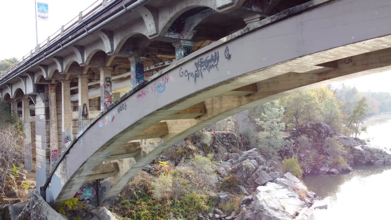 Smooth drone shot, traveling along the side of bridge, a river below and over-grown grass on the other side.