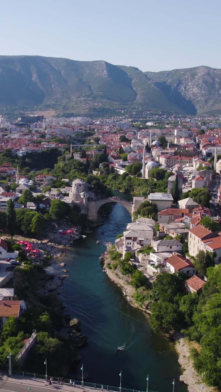 Vertical drone panoramic establishing of Mostar rooftops, river, and historic buildings nestled in midday warm glowing light