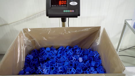 man wearing gloves tosses blue plastic clips into large box on scale in a factory