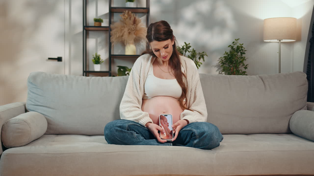 Expecting mother sits on sofa holding smartphone displaying ultrasound image against her pregnant