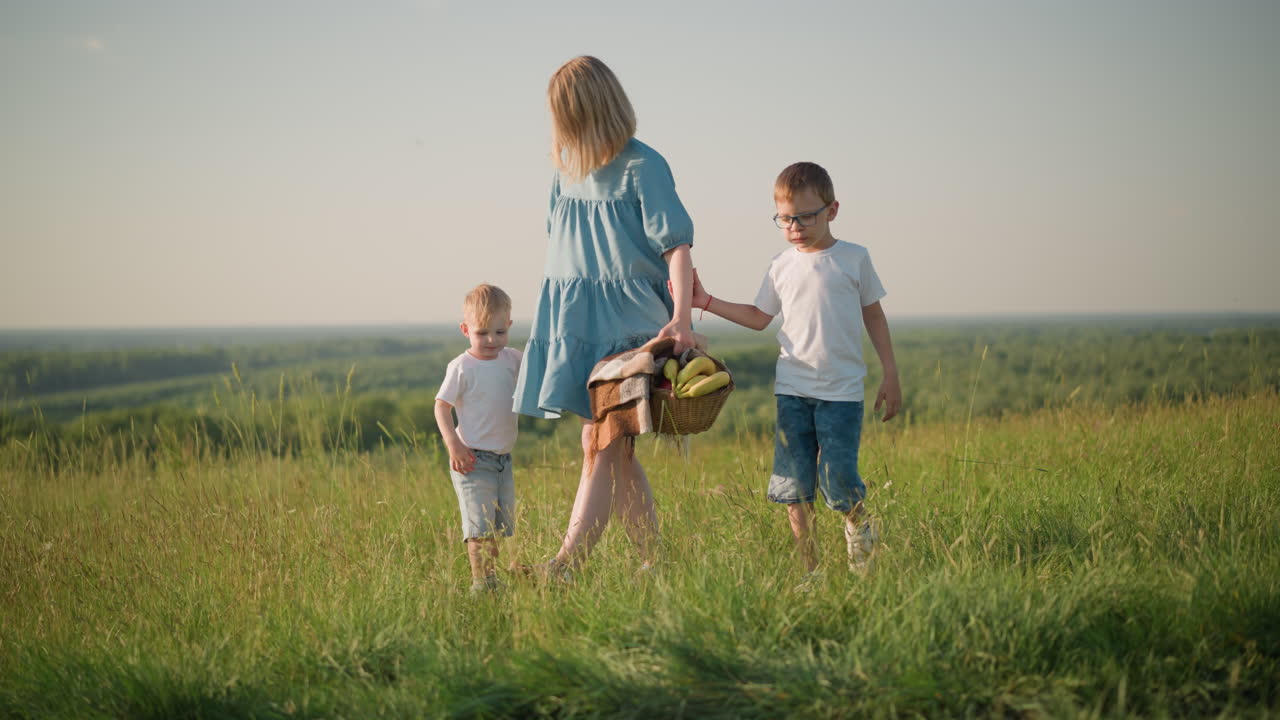 A mother in a blue dress bends down to pick up a basket filled with fresh fruit from the grass while holding hands with her two young sons, both in white tops,as they walk through a sunny, grass field