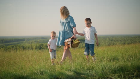 a mother in a blue dress bends down to pick up a basket filled with fresh fruit from the grass while holding hands with her two young sons, both in white tops,as they walk through a sunny, grass field
