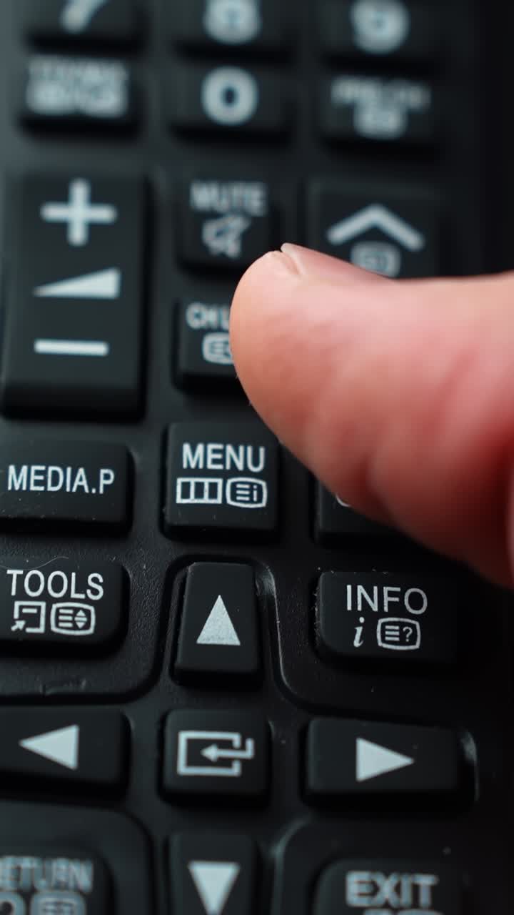 Makrofoto af en finger, der trykker på op-pilen på en TV-fjernbetjening nær knapperne for lydløs og kanaloversigt, lav dybdeskarphed og blødt indendørs lys for et stilrent øjeblik af teknologisk navigation