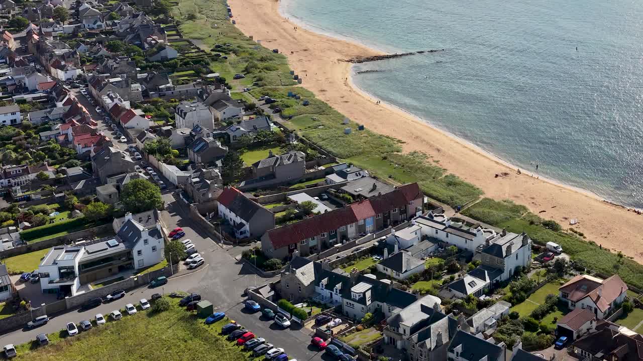 Drone pans above seaside town, sandy beach, and golf course in bright daylight, Fife, Scotland
