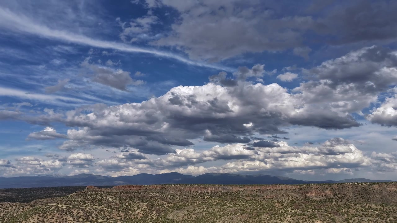 Un timelapse panoramico con drone che cattura nuvole dinamiche su un aspro canyon del sud-ovest. I cambiamenti atmosferici di luce e ombra si evolvono magnificamente attraverso la maestosa cresta desertica e le montagne distanti.