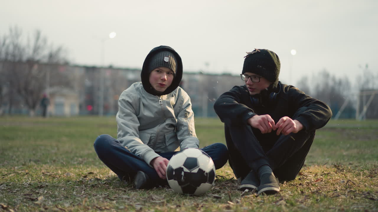 Two boys sitting on a grassy field, engaged in a conversation, the boy wearing glasses holds a stick in his hand, while the other boy in a gray top has a soccer ball in front of him