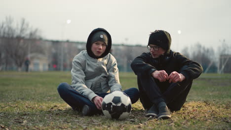 two boys sitting on a grassy field, engaged in a conversation, the boy wearing glasses holds a stick in his hand, while the other boy in a gray top has a soccer ball in front of him