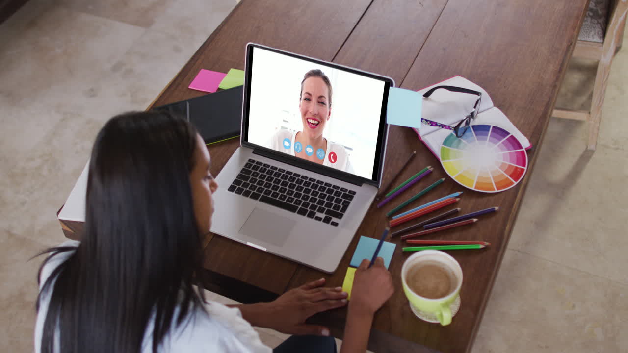 Mujer caucásica usando una laptop en videollamada con una colega, tomando notas