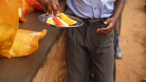 rural african school pupil steps forward to receive fresh cut fruit in tin plate for lunch