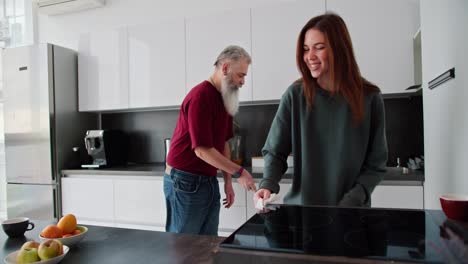 A-man-with-gray-hair-with-a-lush-gray-beard-in-a-red-shirt-together-with-his-adult-brunette-daughter-in-a-green-sweater-is-cleaning-in-a-modern-kitchen