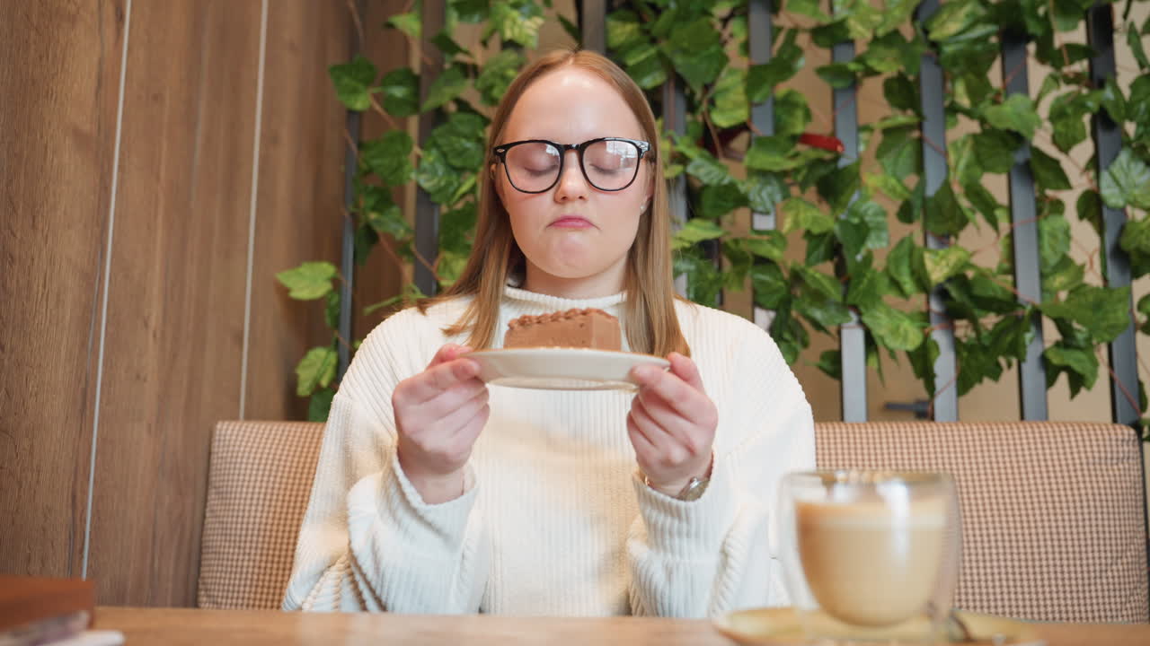 Young woman in glasses holds plate of chocolate cake close to face, smiling as she smells dessert with latte placed on wooden table, surrounded by cozy interior and decorative green plants