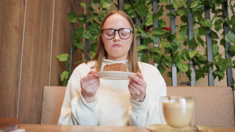 young woman in glasses holds plate of chocolate cake close to face, smiling as she smells dessert with latte placed on wooden table, surrounded by cozy interior and decorative green plants