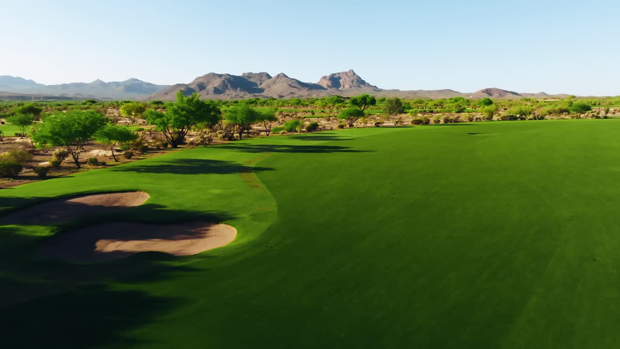 Sweeping drone shot over a wide desert golf fairway with bunkers and distant mountain peaks in view