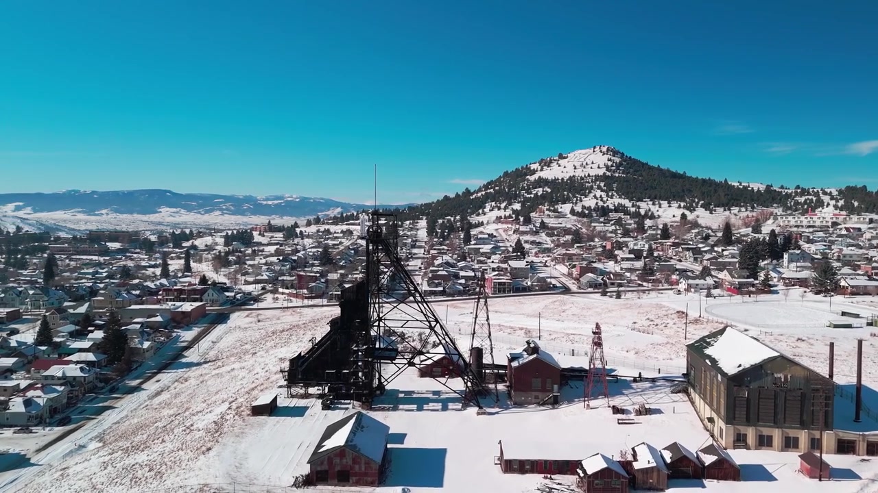 Sorvolo aereo invernale di Butte, Montana, che cattura i castelli minerari storici coperti di neve e la disposizione urbana industriale compatta annidata contro drammatici orizzonti montuosi innevati.