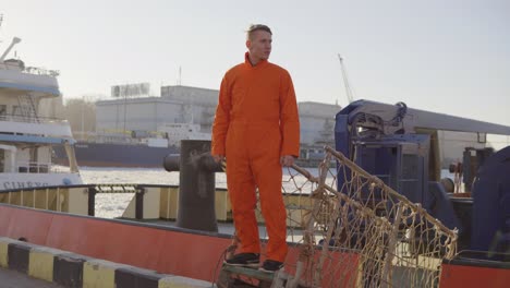 joven trabajador en uniforme posando en el puente de un barco, a punto de atracar en un puerto de la ciudad