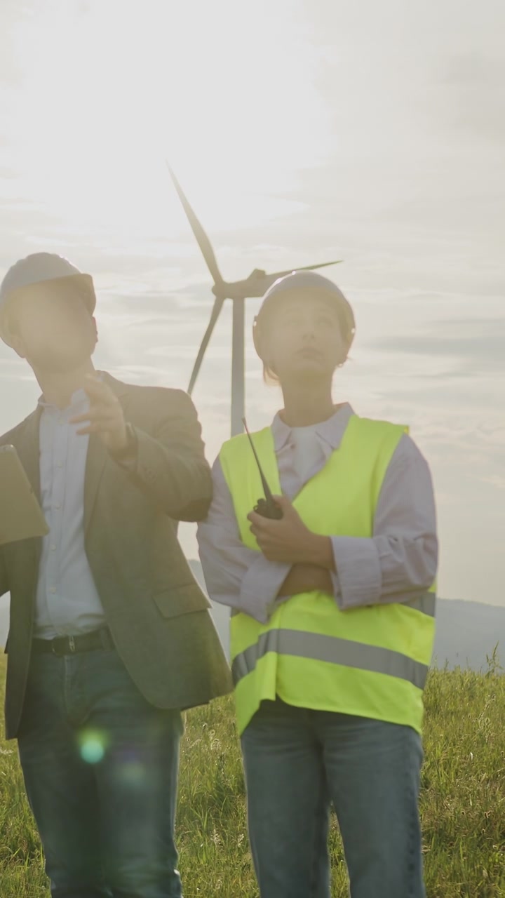 Engineers in safety vests and hard hats stand in a field with wind turbines reviewing data on a