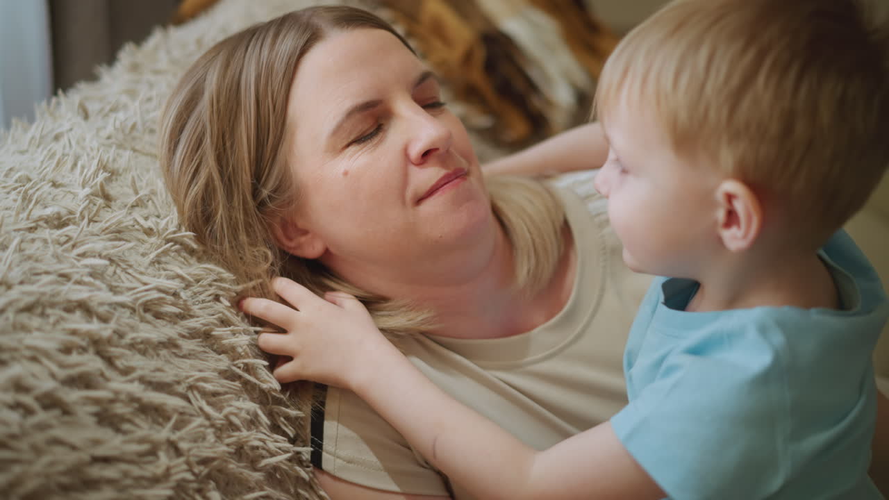 Eine Mutter liegt auf einem weichen Teppich und lächelt warmherzig, während ihr junger Sohn im blauen Hemd sie liebevoll ansieht, ein inniger Moment der Verbundenheit, der Zärtlichkeit, Zuneigung, Verbindung und Glück in einer gemütlichen Wohnatmosphäre zeigt.