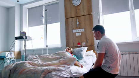 pregnant woman lies in the hospital bed covered with duvet. husband sits beside his wife talking and smiling to her. low angle view.