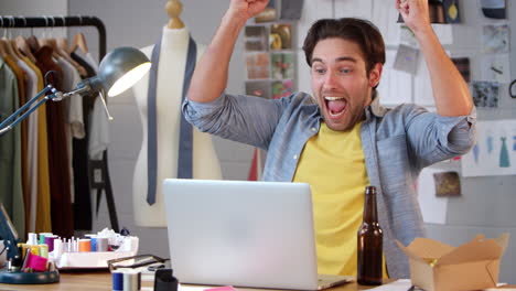 diseñador de moda masculino con una botella de cerveza viendo deportes en la computadora portátil y celebrando en el estudio