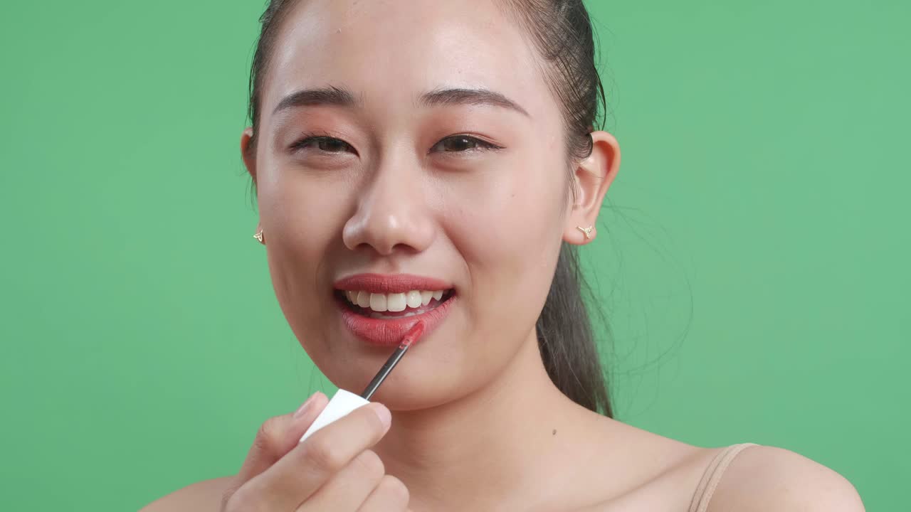 close up de una mujer asiática aplicando lápiz labial y luego sonriendo mientras está de pie en el fondo de la pantalla verde en el estudio