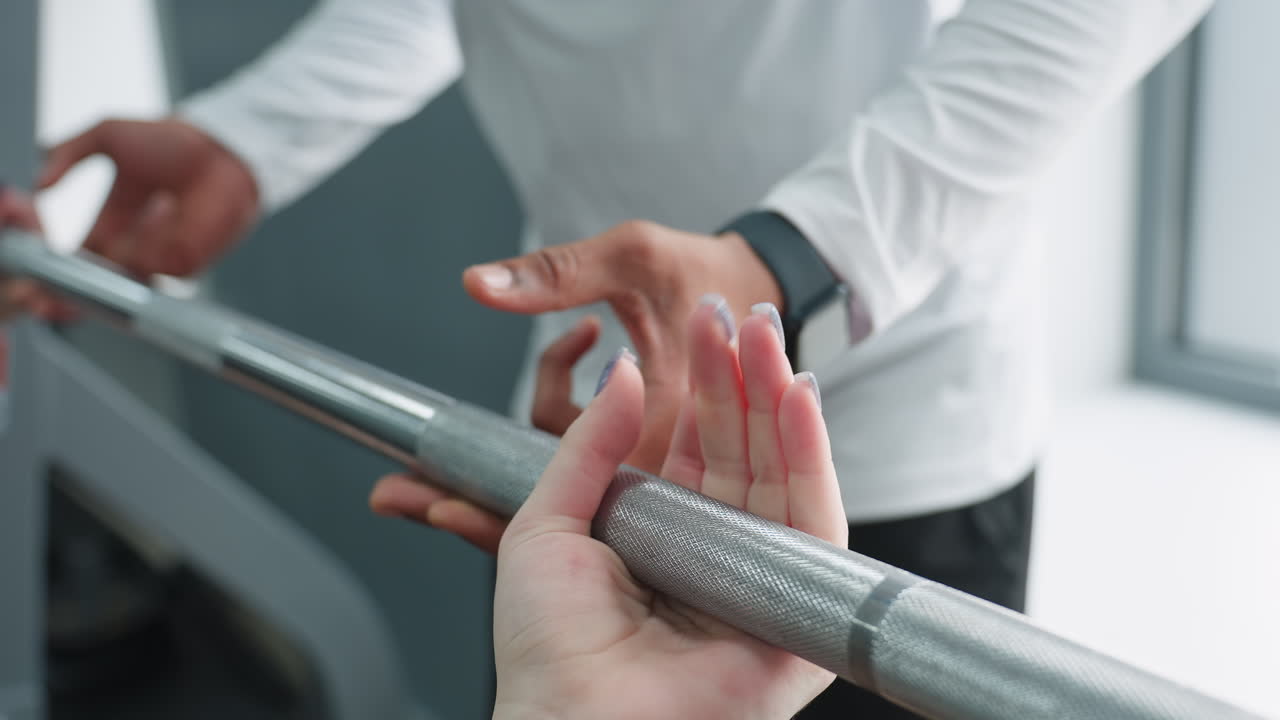 hand view of woman with glittery polished nails gripping barbell during fitness session as male trainer assists from side, focusing on grip technique and teamwork in modern gym with bright lighting
