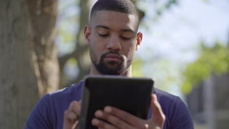 afro-american muscular man on bench in park typing on tablet