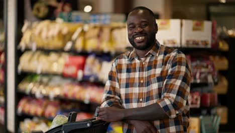 a-man-with-Black-skin-color-in-a-plaid-shirt-stands-near-a-cart-in-a-supermarket,-poses,-looks-at-the-camera-and-smiles