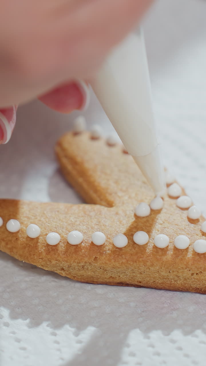 primer plano de la mano decorando una galleta en forma de corazón con glaseado blanco usando una bolsa de tubería, puntos de glaseado intrincados cuidadosamente colocados en una galleta de color marrón dorado descansando en una toalla de papel con textura bajo una iluminación suave