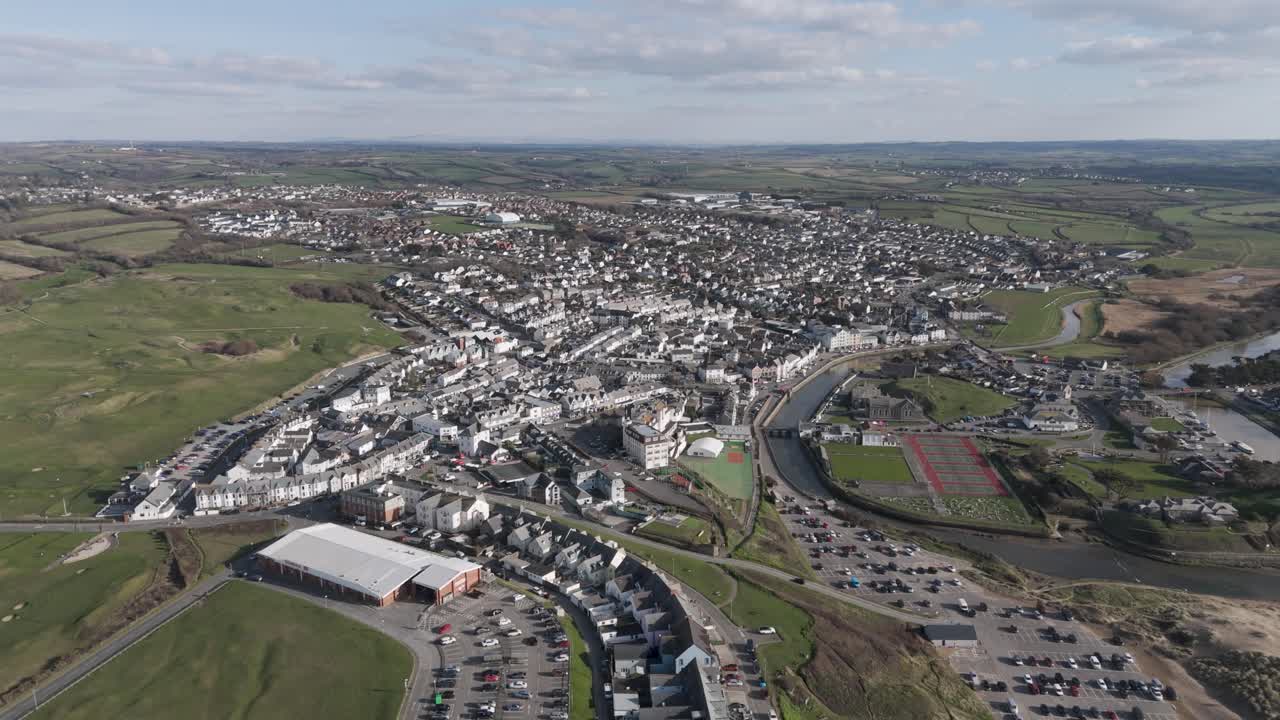 Broad aerial of seaside town where Bude’s rooftops, sports fields and river estuary meet sandy beach and Atlantic surf, blending civic life with coastal recreation and natural scenery