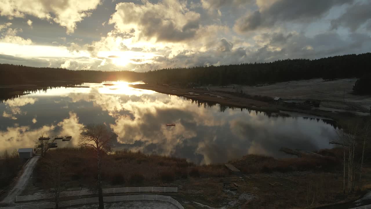 magnífica escena aérea del lago vislumbrado durante la puesta de sol a la luz de la hora dorada, en un día nublado, hermosos reflejos del cielo en el agua, vuelo inverso sobre la tierra del rancho con majestuosos abetos