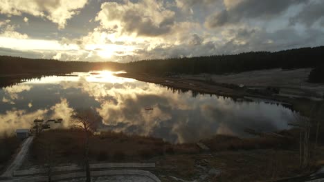 magnificent aerial scene of glimpse lake during sunset in golden hour light, on a cloudy day, beautiful reflections of the sky in the water, reverse flight over ranch land with majestic spruce trees