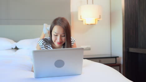 pretty asian woman laying on bed while working on laptop computer