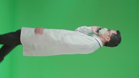 The Side View Of Asian Doctor With Stethoscope Wearing Mask, Glasses, And White Coat Talking On Mobile Phone While Walking On Green Screen Chroma Key.