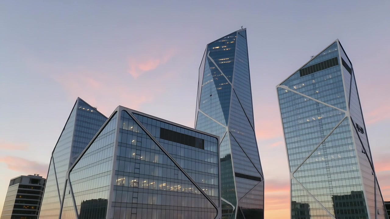 Três torres de vidro refletindo, banhando o pódio no brilho do pôr do sol, de uma vista do telhado, enquanto o crepúsculo se aprofunda.