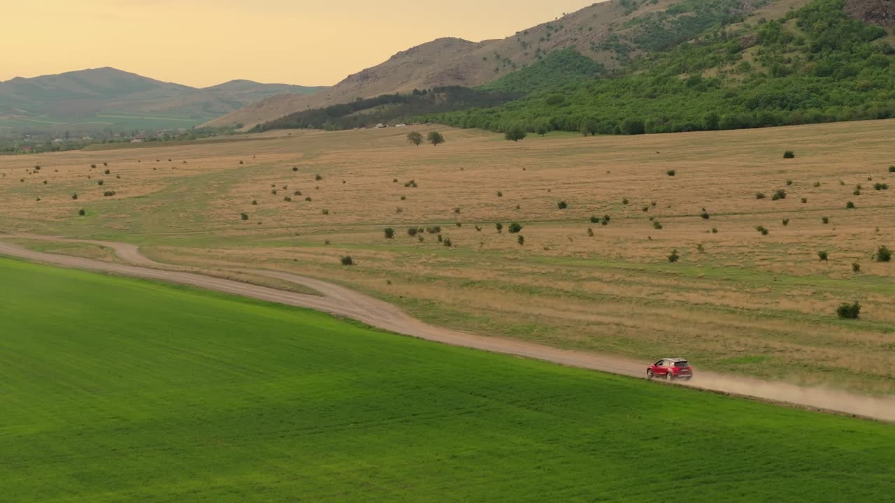 seguimiento aéreo de un coche suv rojo conduciendo en un camino de tierra del país entre campos agrícolas verdes, montañas en el fondo